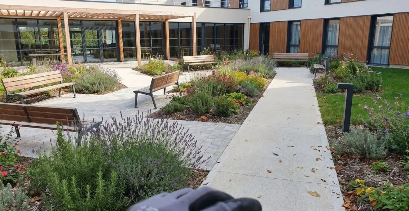 Jardin extérieur d'un établissement pour seniors avec allées accessibles, bancs, parterres fleuris et terrasse couverte en arrière-plan